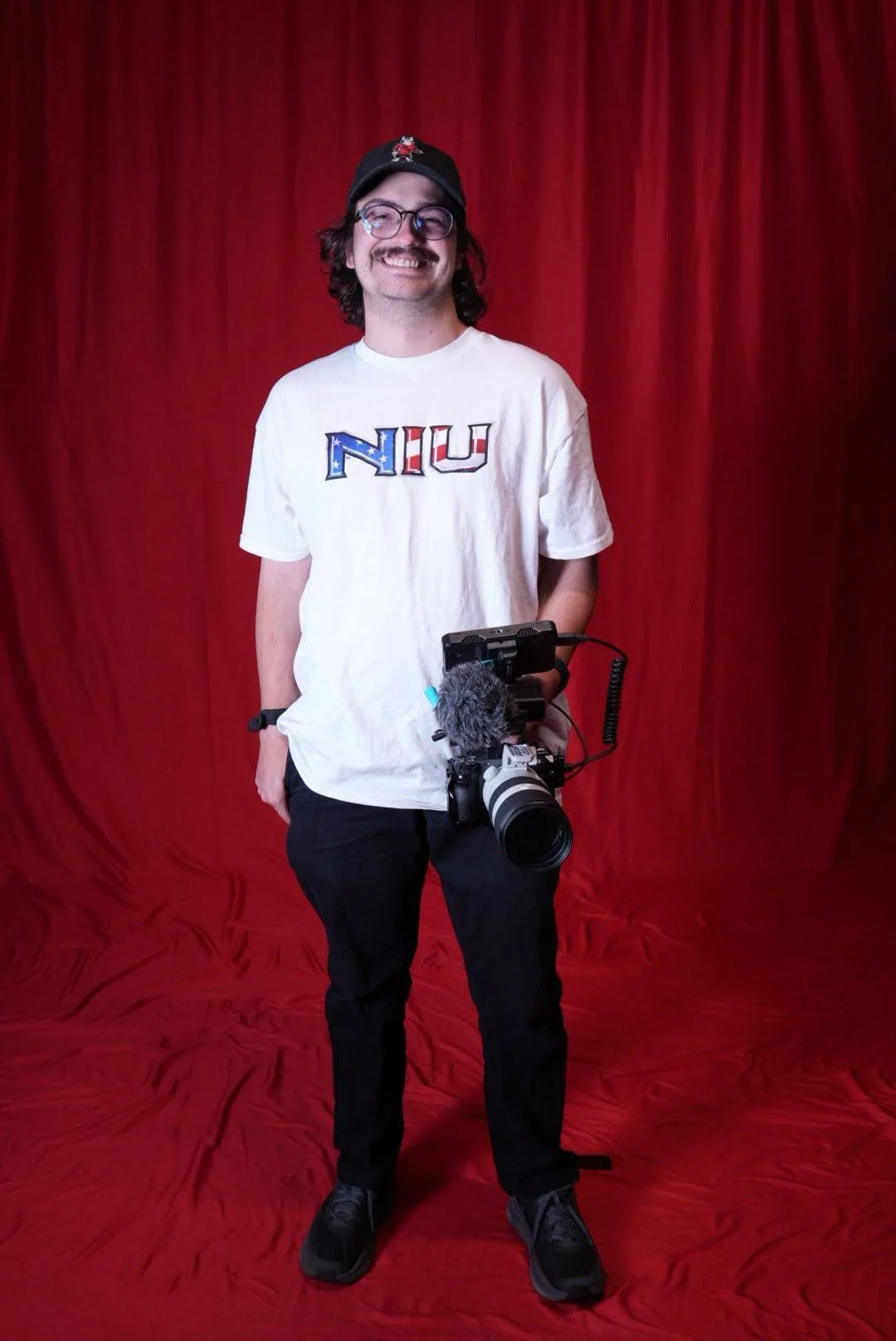 Eric McGill smiling in front of a red backdrop, holding a camera rig with a shotgun microphone